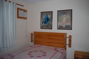 A bedroom with a wooden headboard, a bed with a floral bedspread, two framed paintings on the wall, a window with white curtains, and a small wooden nightstand.