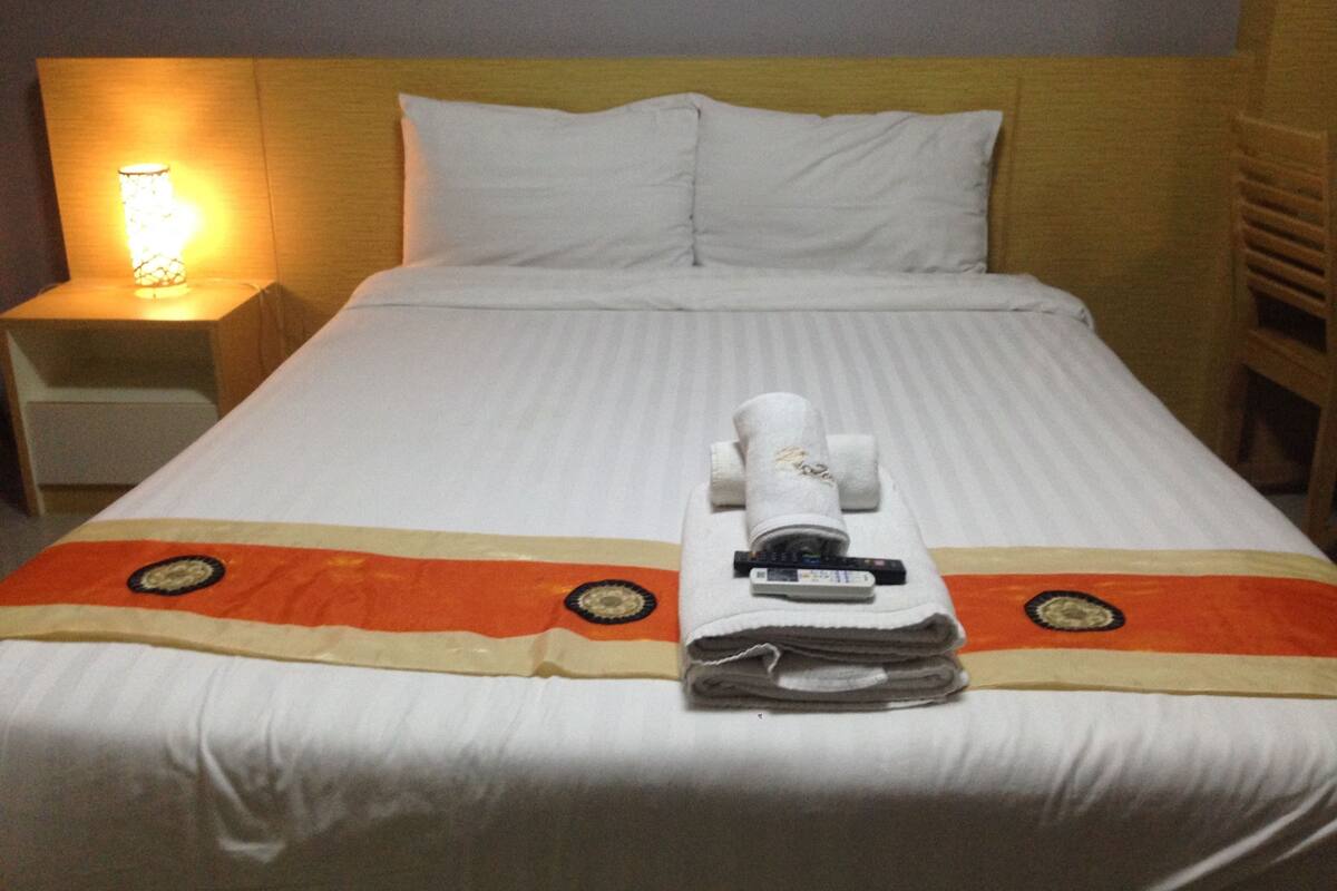 A neatly made bed with white linens, a folded towel, and a remote control on the bed.