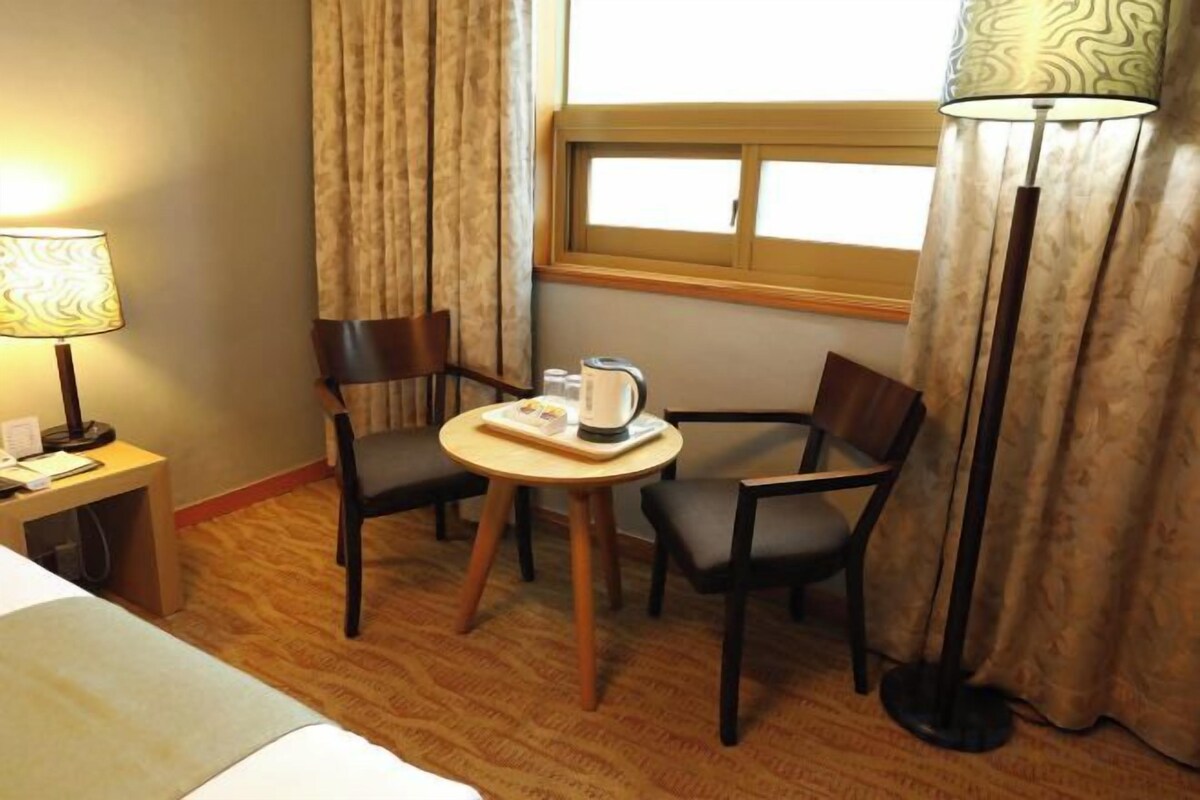 Une chambre d’hôtel avec un lit, un bureau avec une lampe, une table avec des chaises et une fenêtre avec des rideaux.