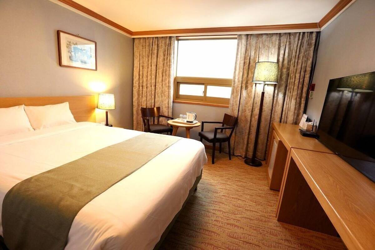 Habitación de hotel con una cama grande, un escritorio, una silla, un televisor, una lámpara y una ventana con cortinas.