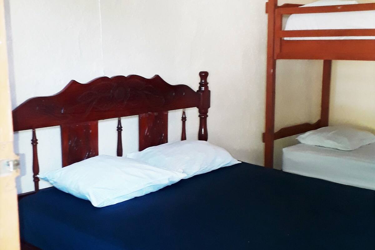Una habitación con una cama de madera, una litera inferior, un ventilador montado en la pared y baldosas de suelo estampadas.