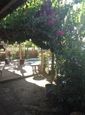 Area patio esterna con piscina, mobili in legno e vegetazione lussureggiante.
