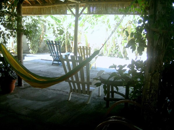 Uma rede sob uma pérgola de madeira, cercada por plantas em vasos e móveis de exterior.