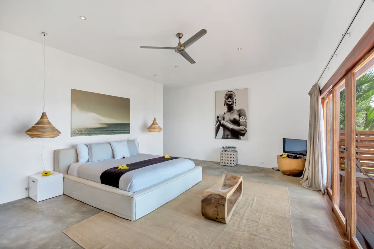 A modern bedroom with a large bed, ceiling fan, artwork, and a TV.