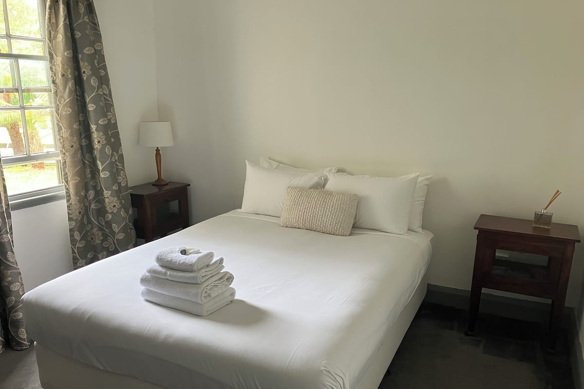 A neatly made bed with white linens and a folded towel stack, a bedside table with a lamp, and a small nightstand with a glass container.