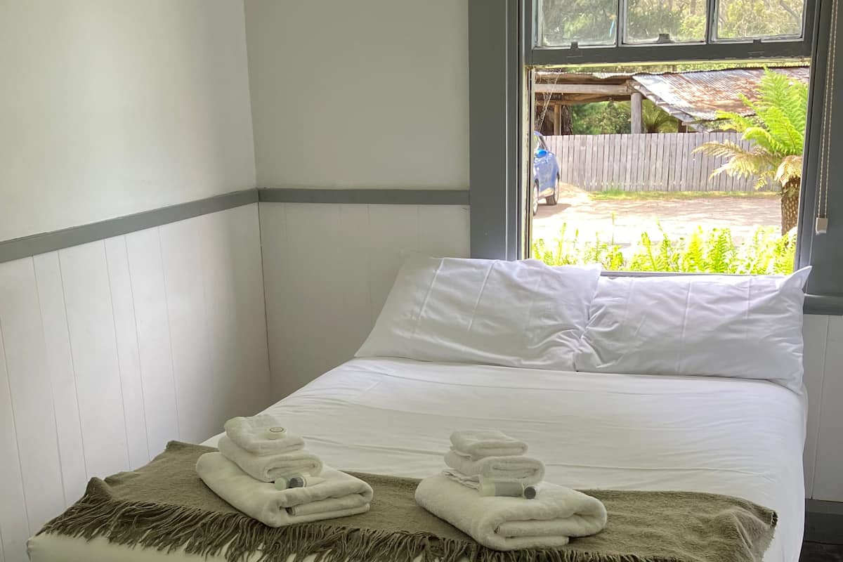 A neatly made bed with white linens and a beige throw blanket, a window with a view of greenery, and a wooden bench in the corner.