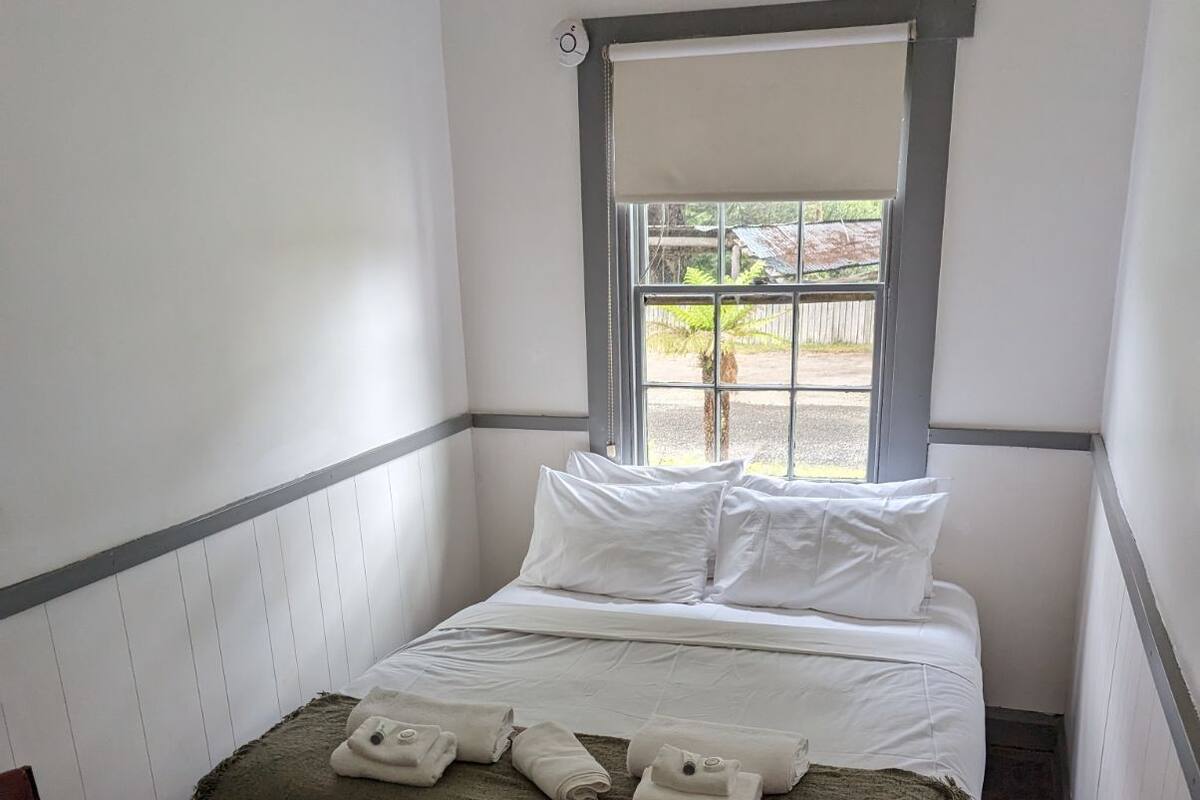 A single bed with white linens, a green throw blanket, and rolled towels. A window with a view of the outdoors is visible.