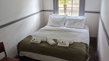 Una cama individual con ropa blanca de cama, una manta verde y toallas enrolladas. Se ve una ventana con vistas al exterior.