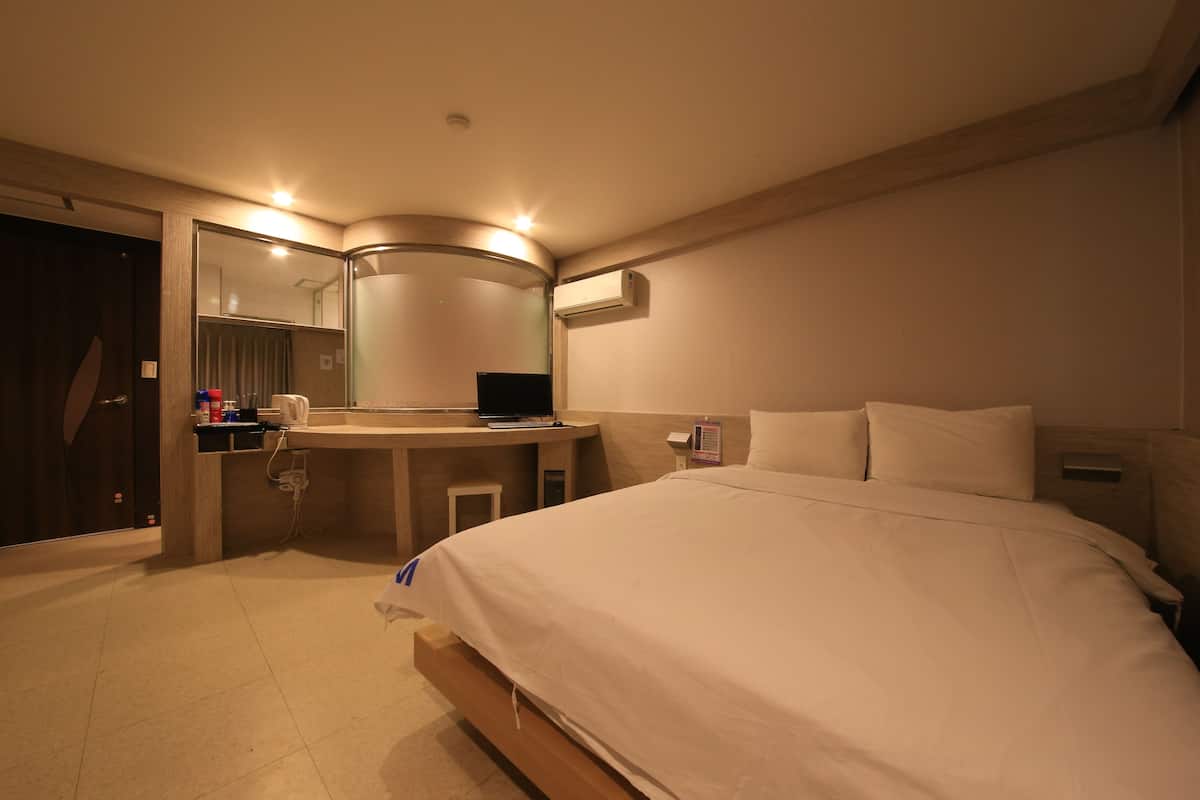 A hotel room with a bed, a desk with a computer, a mirror, and an air conditioning unit.