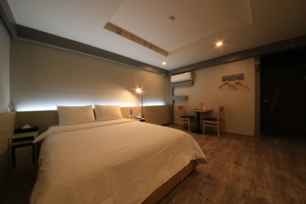 A hotel room with a large bed, a desk with a chair, a small table, and a wall-mounted air conditioner.