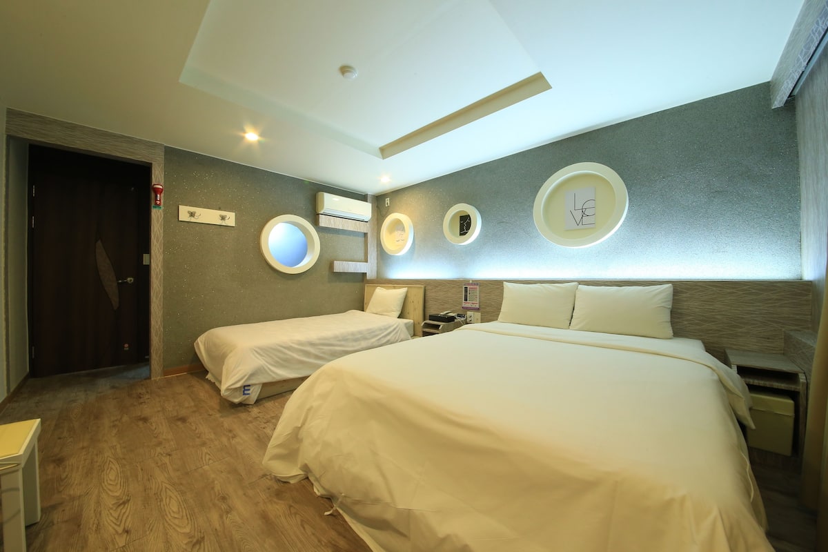 A hotel room with two beds, a wooden floor, a ceiling light, and a wall-mounted air conditioner.
