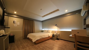 Una habitación de hotel con suelo de madera, una cama con edredón blanco, un escritorio con lámpara y una laptop HP sobre una mesita.