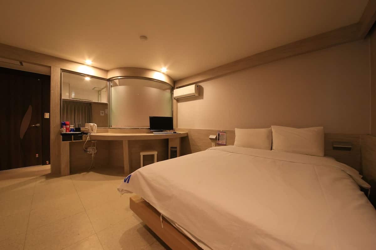 A hotel room with a bed, a desk with a computer, a mirror, and a wall-mounted air conditioner.