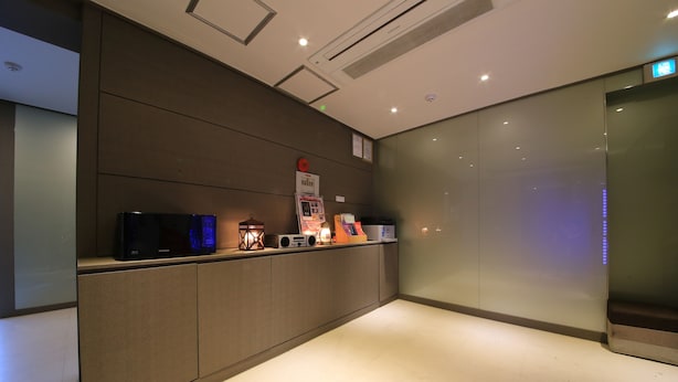 Un área de recepción con mostrador, pantalla digital, una pequeña mesa con lámpara y un letrero en la pared.