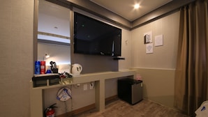 Habitación de hotel con un televisor grande montado en la pared, un escritorio pequeño con un hervidor, un extintor y un cubo de basura.