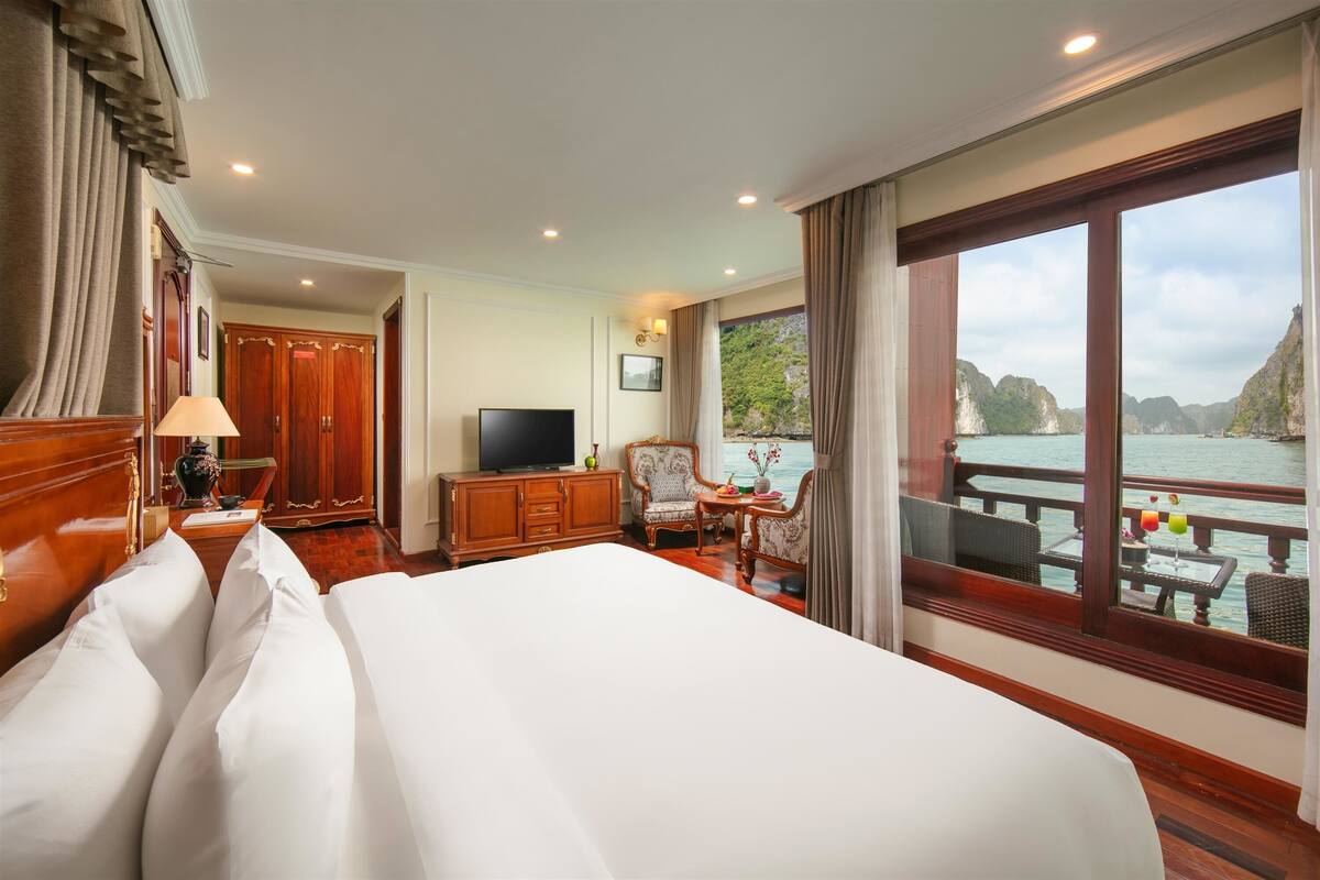 Habitación de hotel con una cama grande, televisión, cómoda de madera, silla, mesita pequeña y balcón con vista al agua.