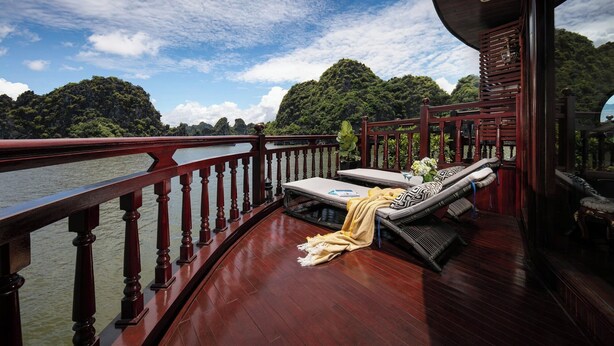 Une terrasse en bois avec une rambarde rouge, un coin salon avec un coussin et une couverture, et une vue sur une rivière et des collines boisées.