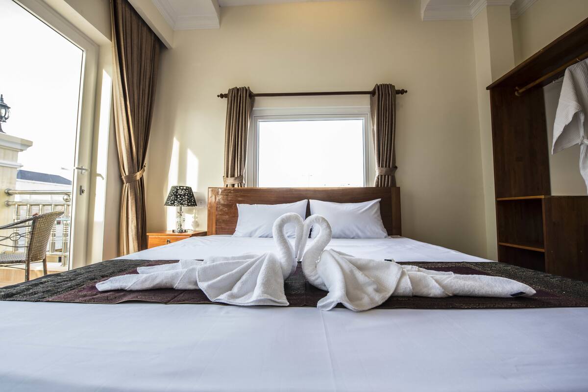 Quarto de hotel com cama, mesa de cabeceira, luminária, janela com cortinas e vista exterior.