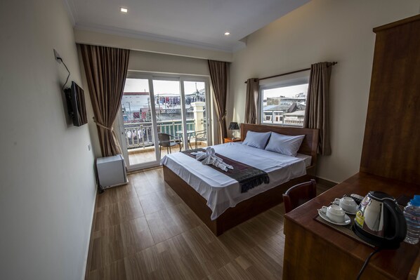 A hotel room with a bed, a desk with a kettle and cups, a TV, and a balcony with a view of buildings. - Phnom Penh Le Vincent Hotel (Phnom Penh)