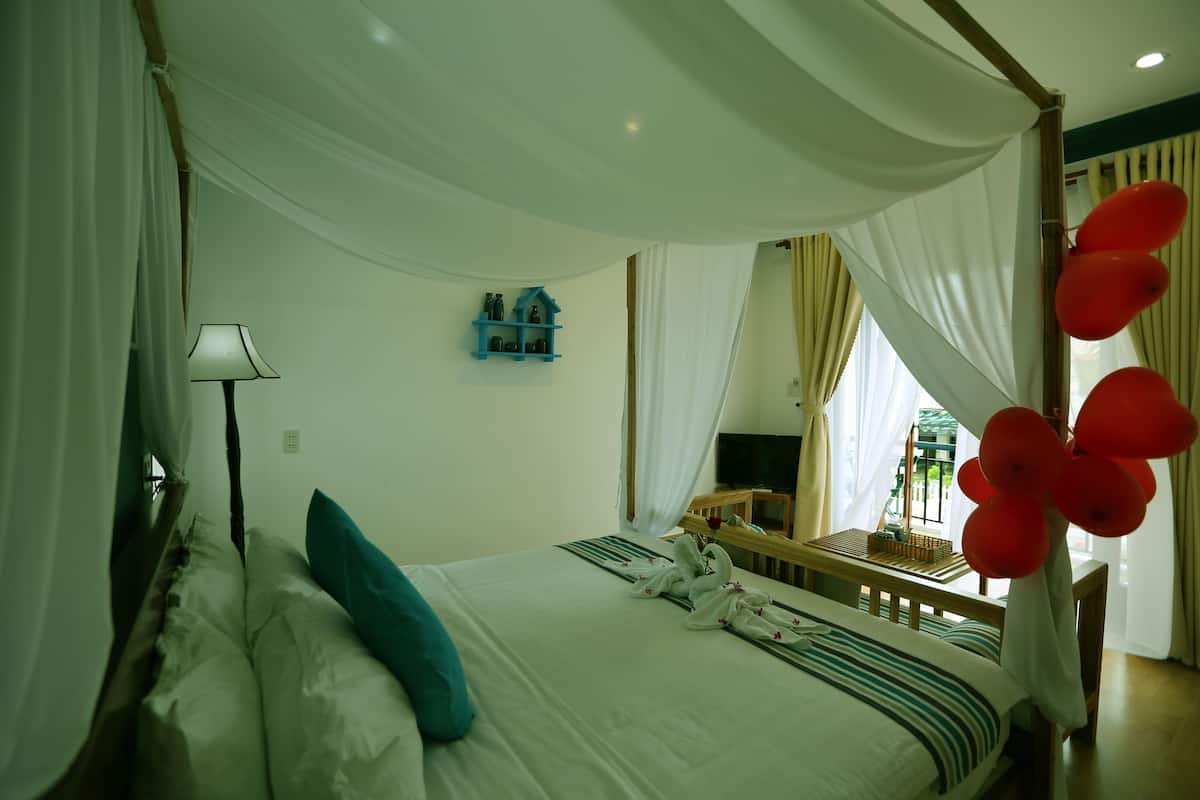 Un dormitorio con cama con dosel, una mesita de noche con lámpara, un televisor y una ventana con cortinas.