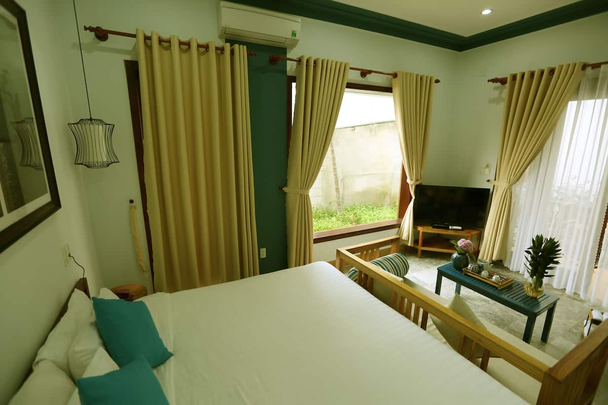 Habitación de hotel con cama, televisión, una mesita con una piña y un jarrón, y balcón con cortinas.