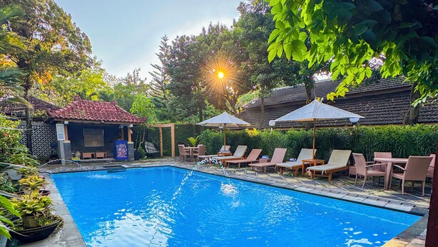 Une piscine entourée de verdure et d’un bâtiment traditionnel, avec des chaises longues et des parasols à proximité.