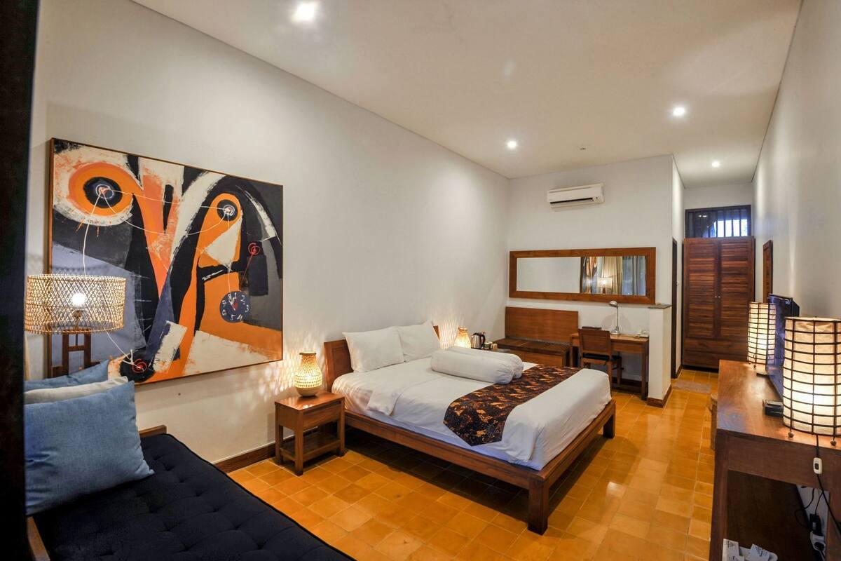 Een hotelkamer met een bed, nachtkastje, lamp, stoel, bureau en een groot abstract schilderij.