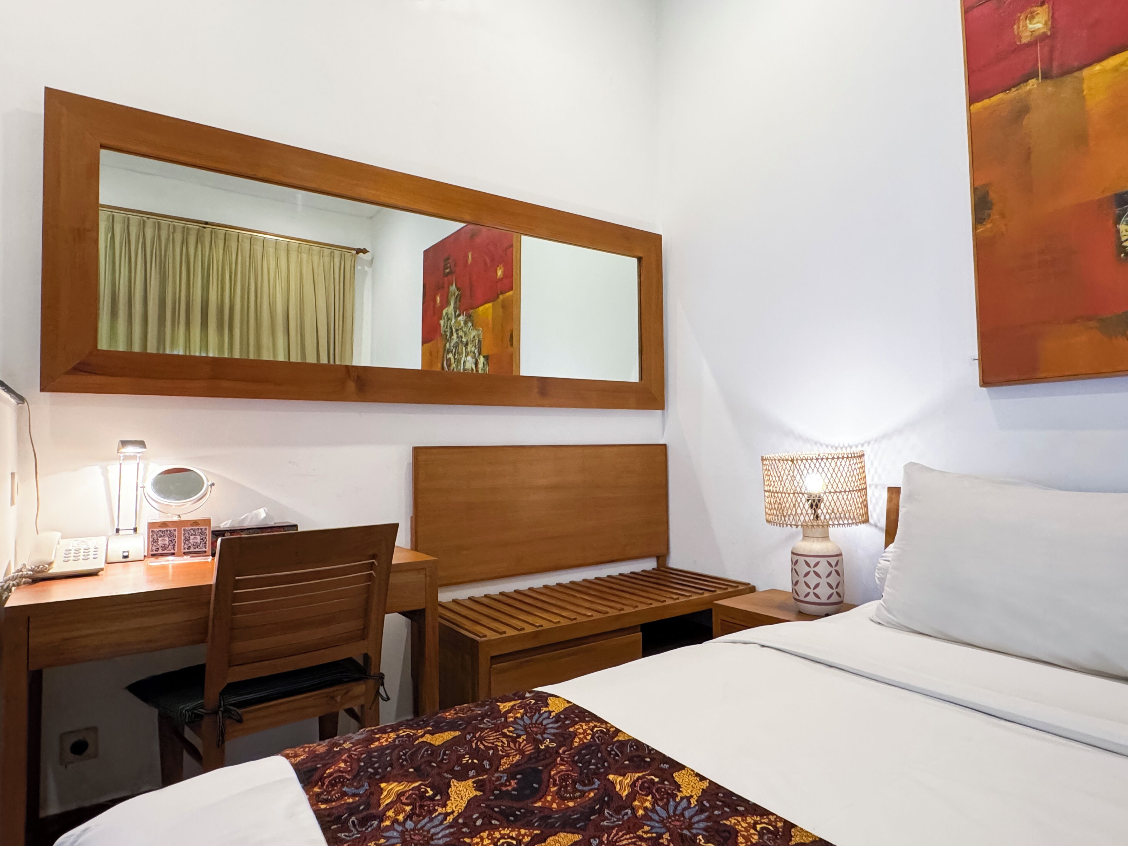 A hotel room with a wooden desk, a chair, a bed with a patterned bedspread, a bedside lamp, a wall-mounted mirror, and a painting.