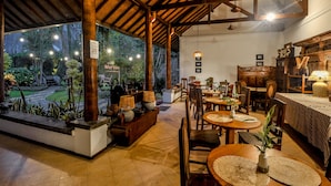 Un restaurant chaleureux avec un plafond en bois, des tables rondes et des chaises. On y trouve des plantes en pot et une vue sur un jardin à l’extérieur.