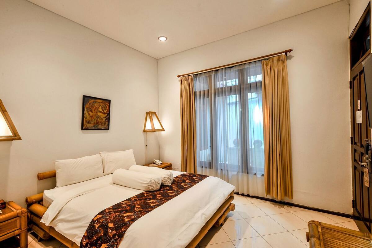 Een hotelkamer met een bed, bedlampjes, een schilderij, een raam met gordijnen en een houten garderobekast.
