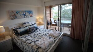A bedroom with a bed, a nightstand, a lamp, a painting, a window with curtains, and a balcony with a table and chairs. - Beachfront Shoal Bay (Nelson Bay)