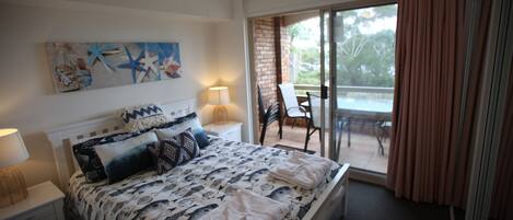 A bedroom with a bed, a nightstand, a lamp, a painting, a window with curtains, and a balcony with a table and chairs.