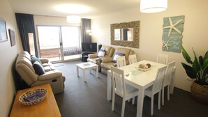 A spacious living area with a sofa, a dining table, and a TV. There is a balcony with a view. - Beachfront Shoal Bay (Nelson Bay)