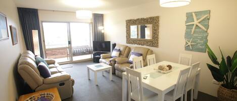 A spacious living area with a sofa, a dining table, and a TV. There is a balcony with a view.