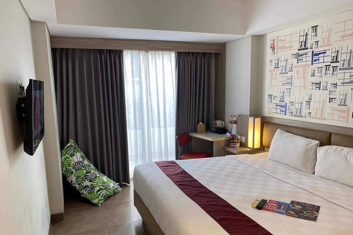 Habitación de hotel con una cama grande, un escritorio con silla, un televisor montado en la pared y una ventana con cortinas.