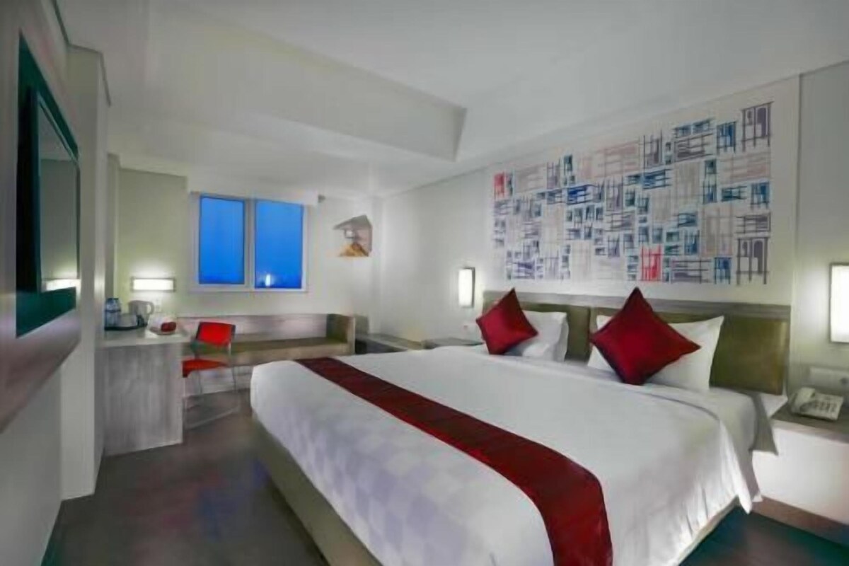 Habitación de hotel con una cama grande, dos mesitas de noche, un escritorio y una silla. Se observan dos ventanas y una pared con un diseño decorativo.