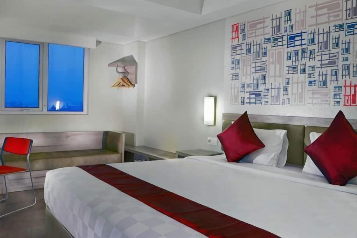 Habitación de hotel con una cama grande, zona de estar, pared con arte abstracto y ventana con vista al cielo azul.