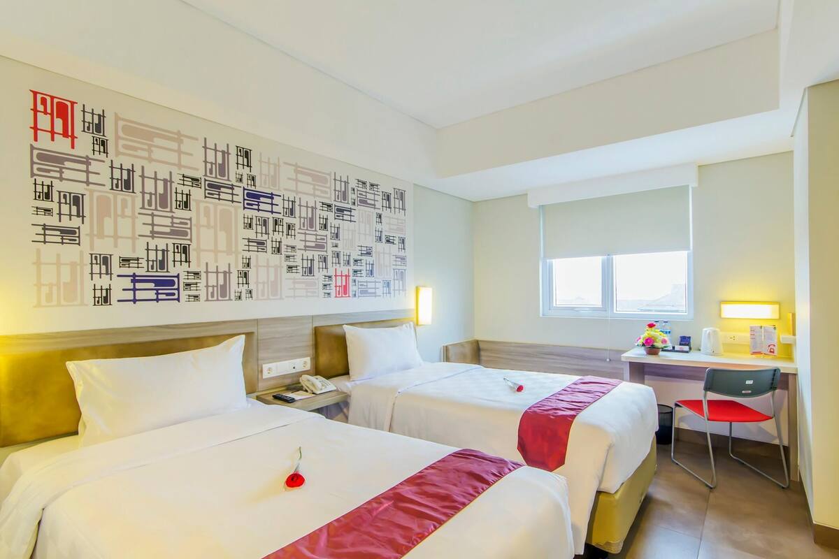 Habitación de hotel con dos camas, un escritorio, una silla, una ventana y una pared decorada con arte abstracto.