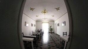 Un couloir avec un sol à carreaux, des murs blancs et un lustre. Des tableaux encadrés ornent les murs, et une plante est placée au centre.
