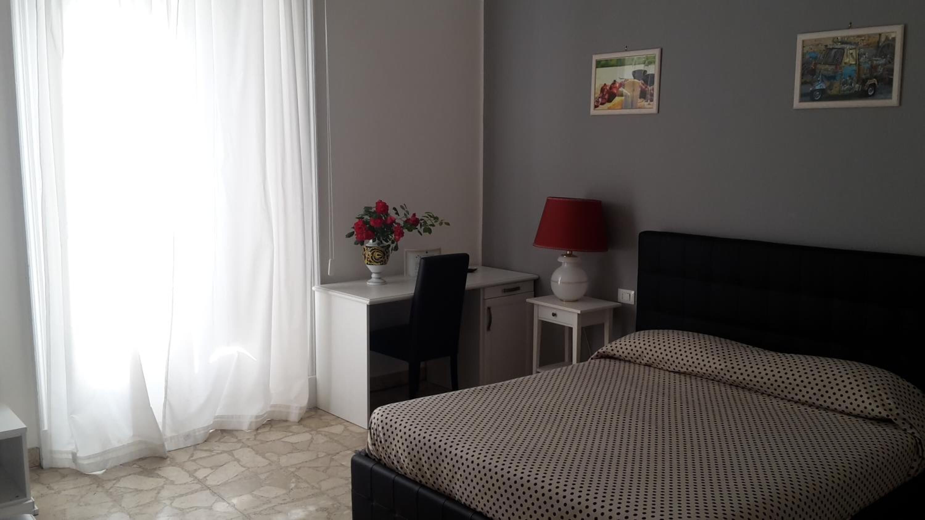 A bedroom with a bed, a desk with a chair, a lamp, and a vase with flowers.