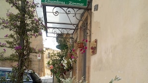 Une rue étroite avec un bâtiment portant l’inscription « GUALTIERO Camere - Rooms » et « Chambres - Zimmer ». Des plantes en pot et une voiture sont garées dans la rue.