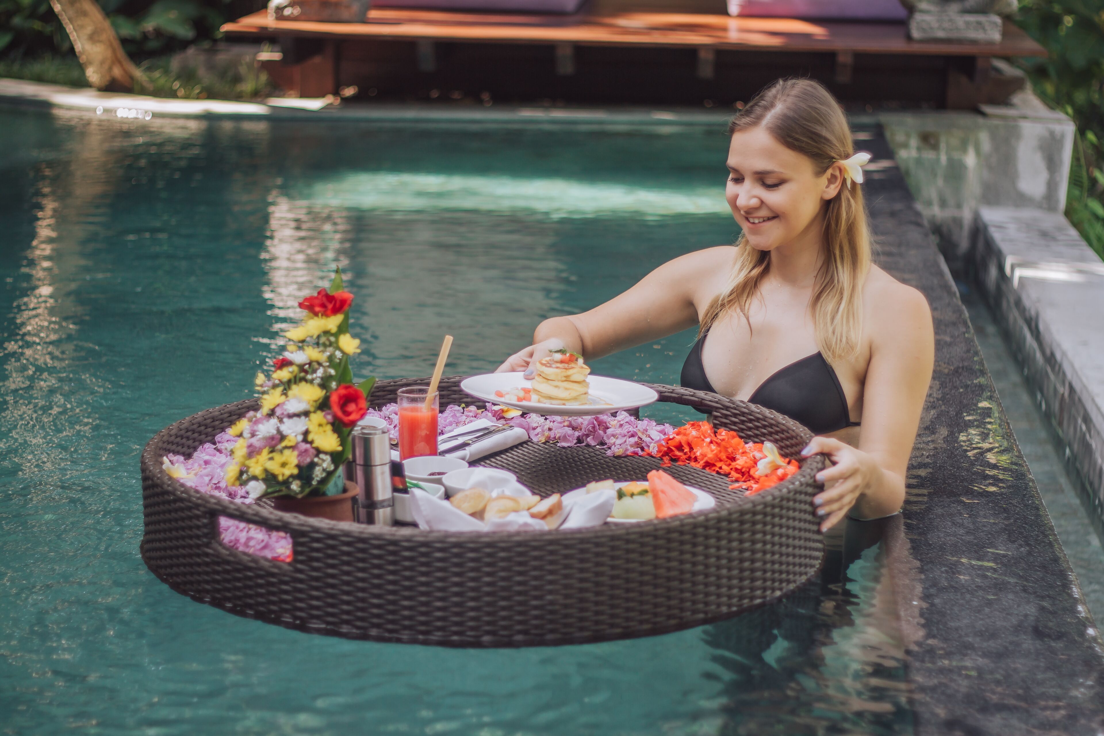 A person holding a wicker tray with food and drinks by a pool.