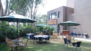 Un patio al aire libre con mesas y sillas redondas, rodeado de follaje y sombrillas.