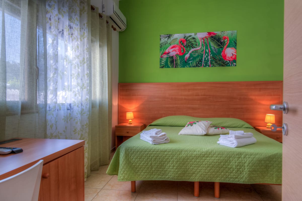 A neatly made bed with green bedding, a desk with a chair, and a painting of flamingos on the wall.