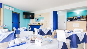 A dining area with tables set for a meal, a kitchen area with a counter and appliances, and a television mounted on the wall. - Aquarium Residence (Manduria)