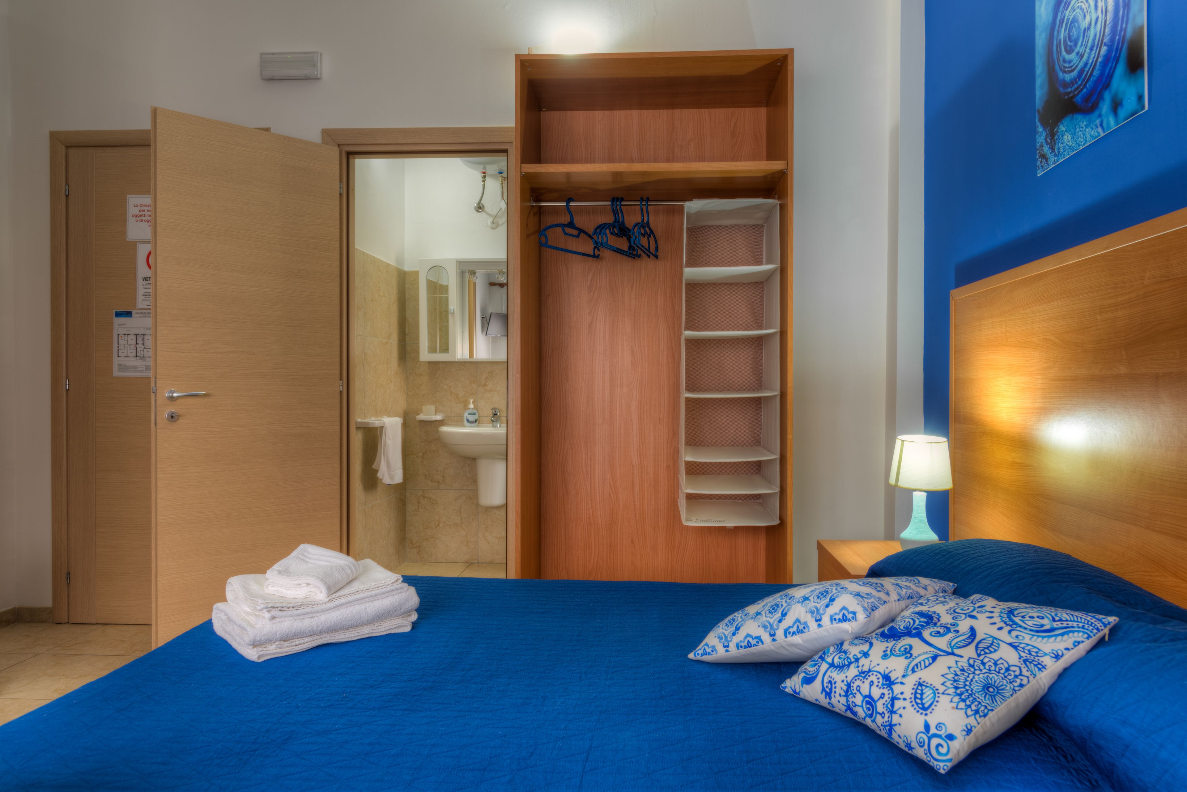 A bedroom with a bed, a wooden wardrobe, a bathroom visible through an open door, and a nightstand with a lamp.
