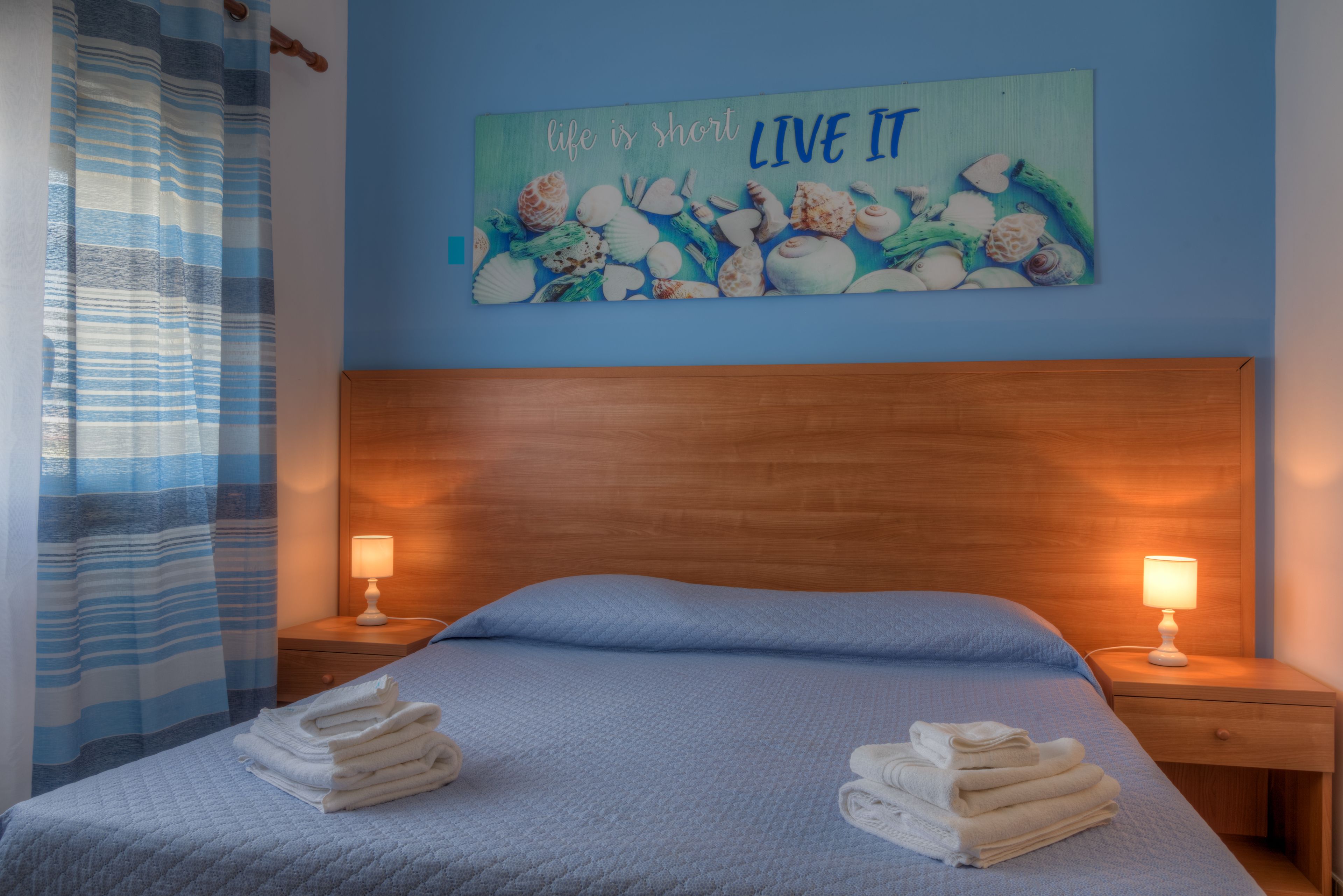 Una cama bien hecha con ropa de cama azul, dos lámparas de noche y una obra de arte en la pared con conchas y la frase "life is short LIVE IT".