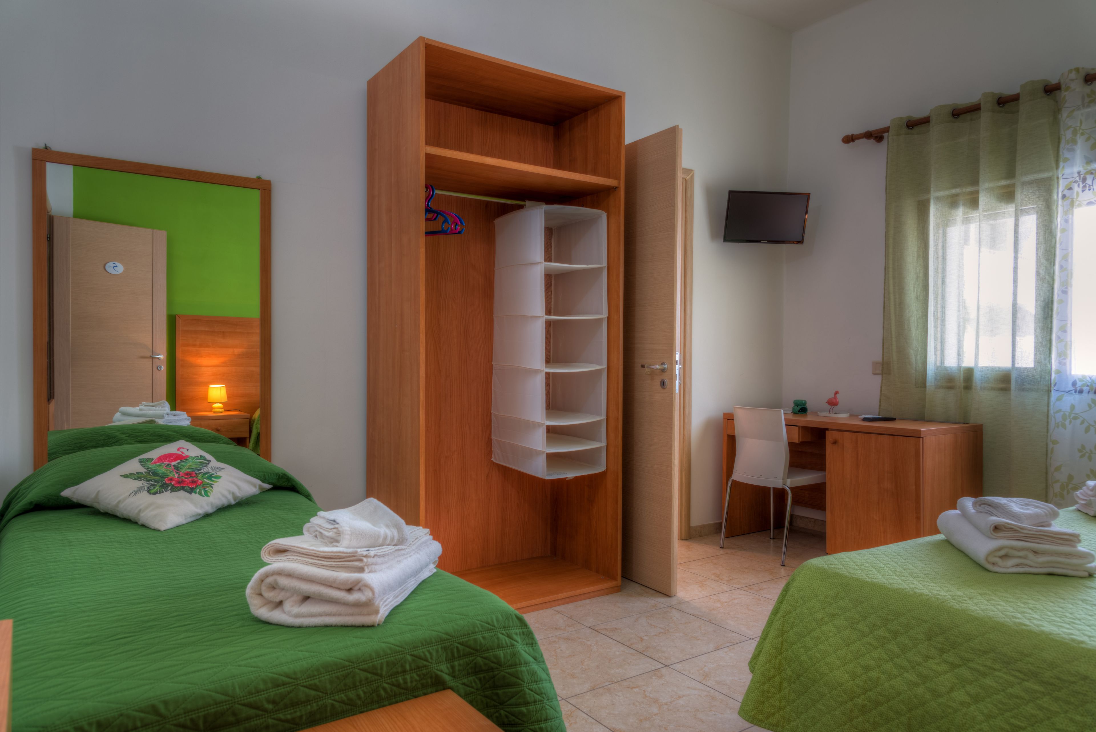 A room with two beds, a wooden wardrobe, a desk, a chair, and a television on the wall.