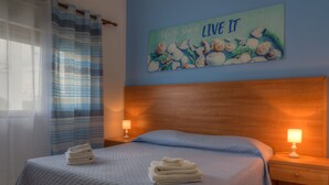 A neatly made bed with folded towels, a blue wall with a 'Live It' artwork, and two bedside lamps. - Aquarium Residence (Manduria)