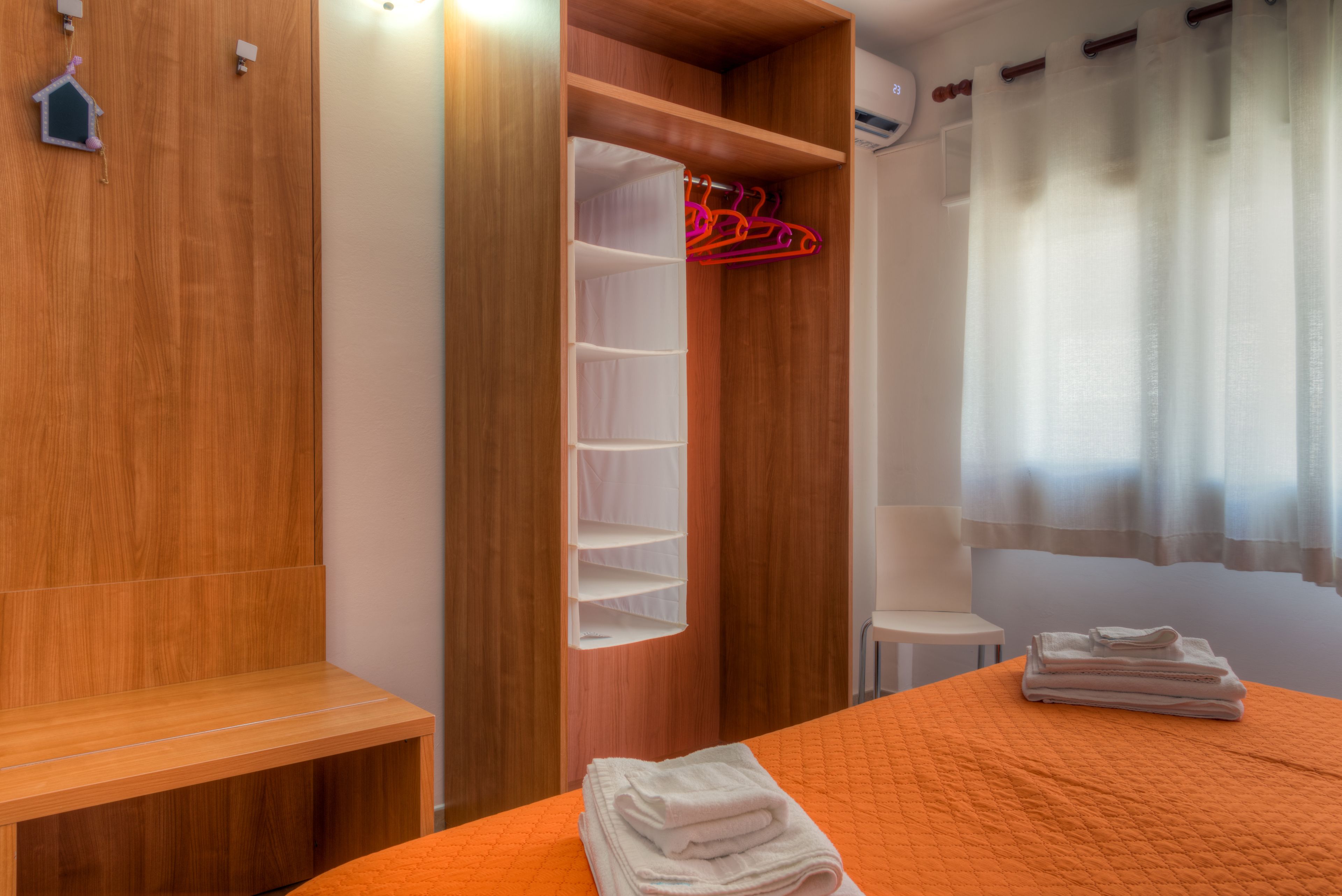 A hotel room with a wooden wardrobe, a bed with an orange bedspread, and a window with sheer curtains.